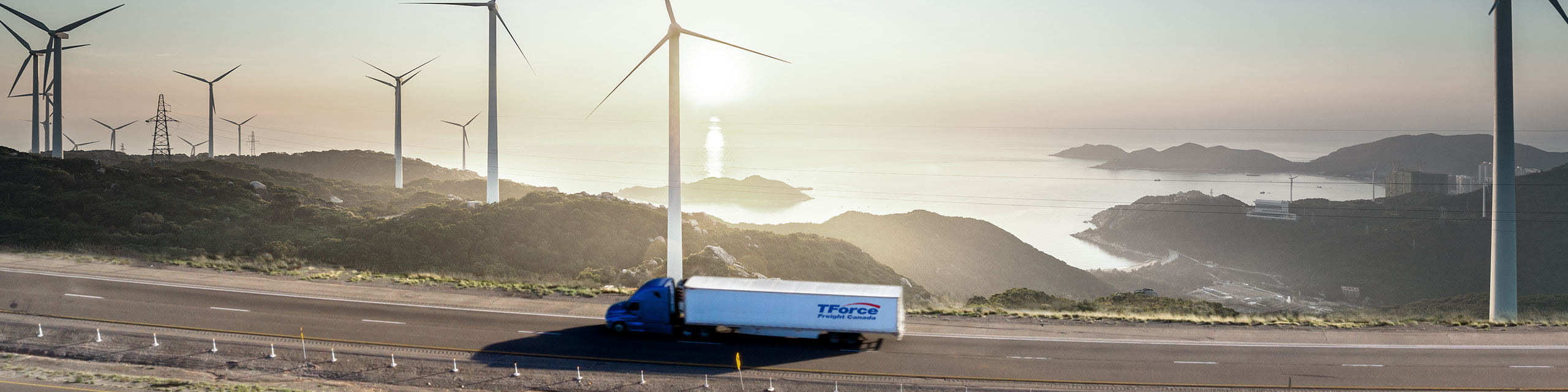 Camion de TForce Freight Canada roulant sur l’autoroute avec cabine bleue devant un lac et un champ d’éoliennes