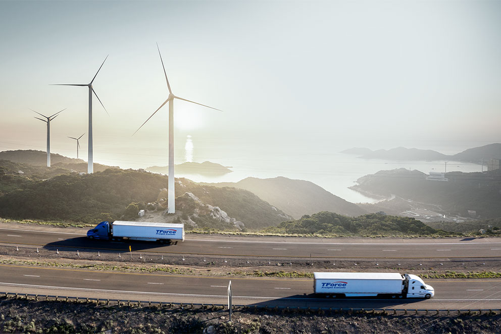 TForce Freight Canada highway truck with blue cab driving in front of lake and windmill field